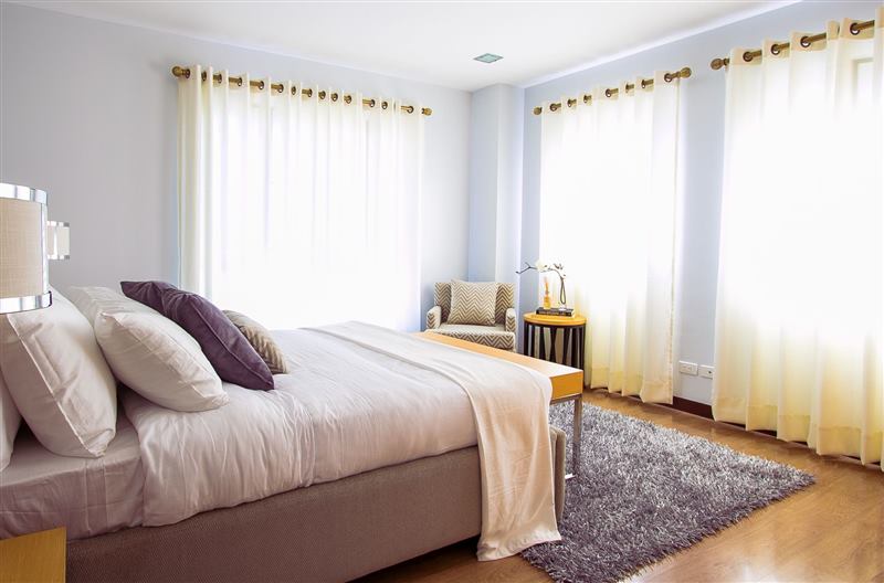 Sunlit bedroom with white linens, sheer curtains, and a chair